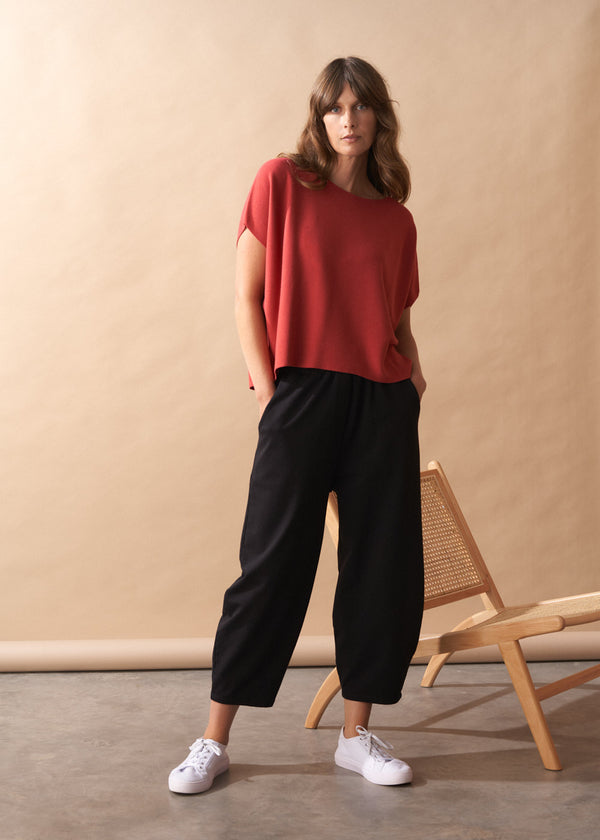 Woman wearing a red short sleeve knitted sweater and black pants standing against a beige wall with a wooden chair.