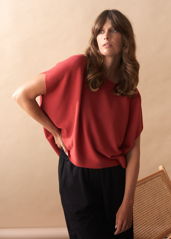 Woman wearing a red short sleeve knitted sweater and black pants against a beige wall.