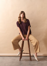 Woman sitting on a wooden stool wearing a burgundy knit top and camel wide leg trousers against a beige wall