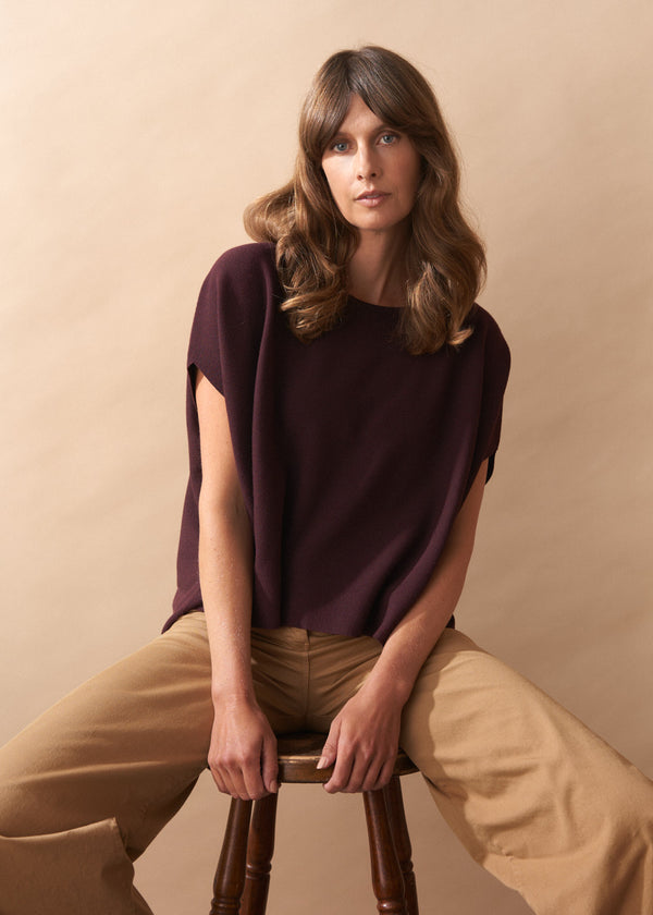 Woman sitting on a stool wearing a burgundy knit top and camel wide leg trousers against a beige background