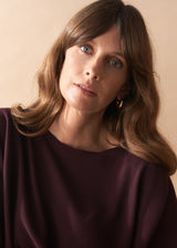 Woman with wavy brown hair wearing a burgundy knit top and camel wide leg trousers against a beige background