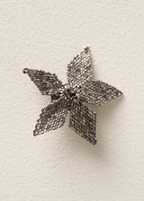 A silver beaded flower-shaped brooch with a textured mesh-like pattern, displayed on a textured off-white background.