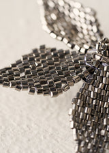 Close-up of a metallic silver beaded decorative object, possibly a butterfly or floral form, made from small rectangular seed beads on a light textured background.