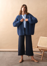 Woman wearing a chunky knit blue cardigan over a light blue shirt with dark pants, standing against a beige wall.