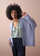 Woman wearing a dusky lavender chunky oversized knit cardigan, over a floral top and blue, draw string trousers.