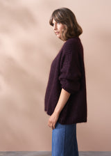 Woman wearing a dark purple chunky knit sweater and blue jeans against a beige background.