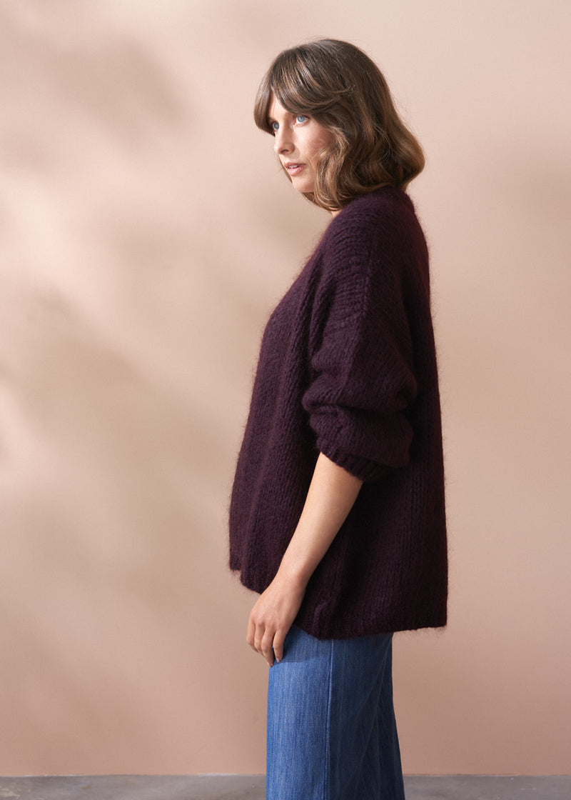 Woman wearing a dark purple chunky knit sweater and blue jeans against a beige background.
