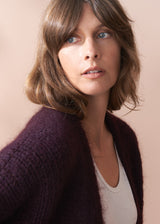 Woman wearing a dark purple chunky knit cardigan against a light pink background.