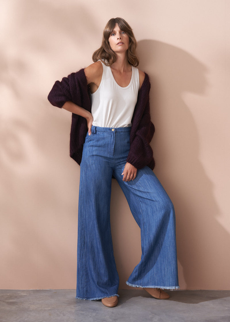 Woman wearing a Dina chunky knit cardigan in dark purple, with white tank top and blue jeans, against a beige wall.
