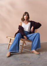 Woman sitting on a wooden chair wearing a white top, blue jeans, and a dark chunky knit cardigan against a beige background.