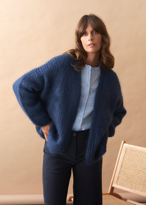 Woman wearing a blue cardigan over a light blue shirt and dark pants against a beige background.