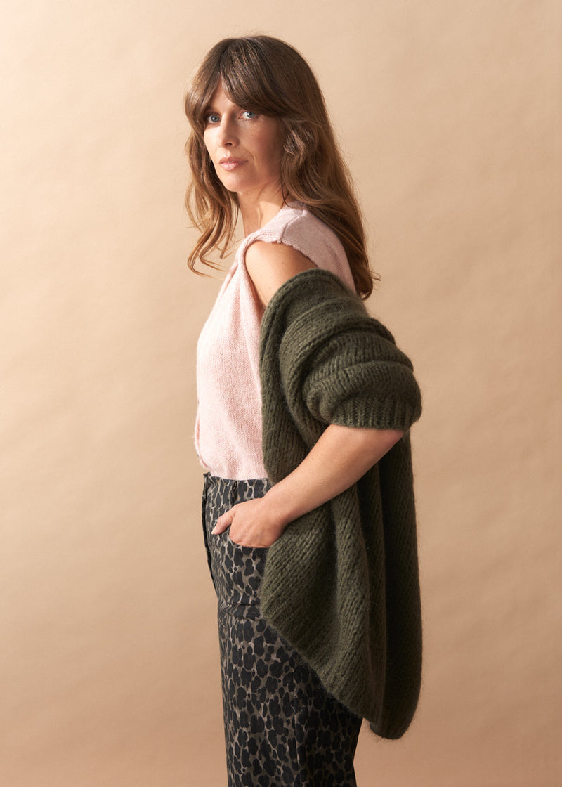 Woman wearing a green cardigan over a pink top and patterned pants against a beige background