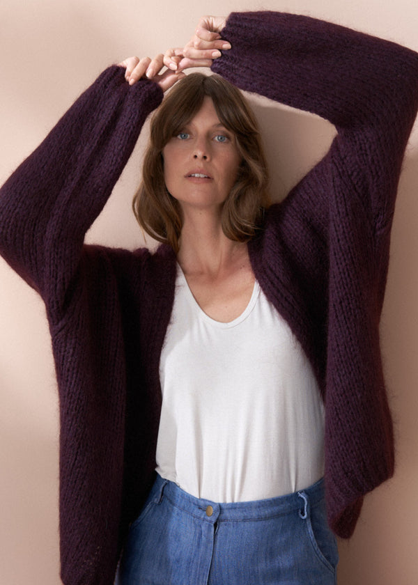 Woman wearing a dark purple cardigan over a white top and blue jeans, holding the cardigan open.