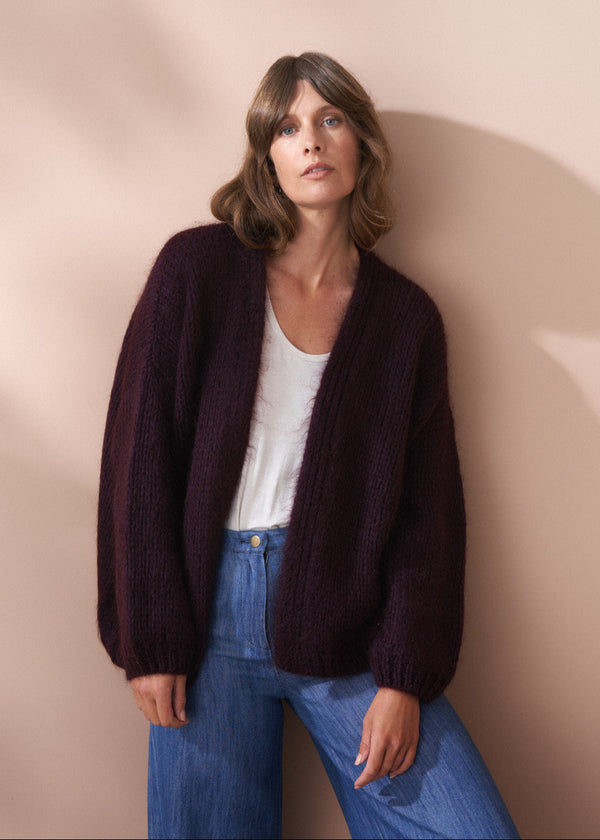Woman wearing a dark purple cardigan over a white shirt and blue jeans against a beige wall.