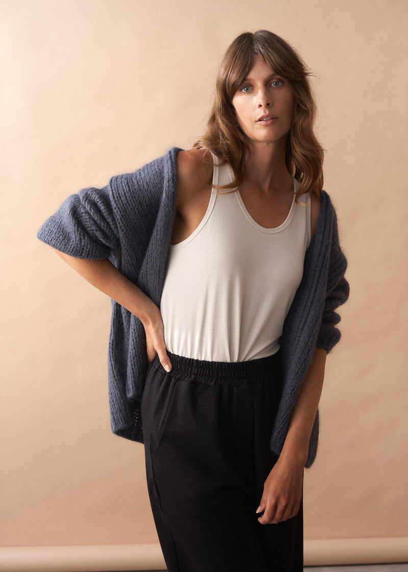 Woman wearing a slate grey chunky knit cardigan, white tank top, and black trousers against a beige background