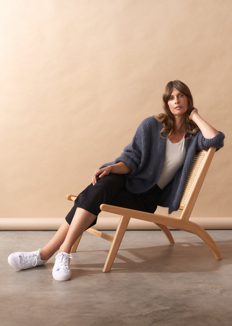 Woman wearing Dina chunky knit cardigan, sitting on a wooden chair against a beige wall