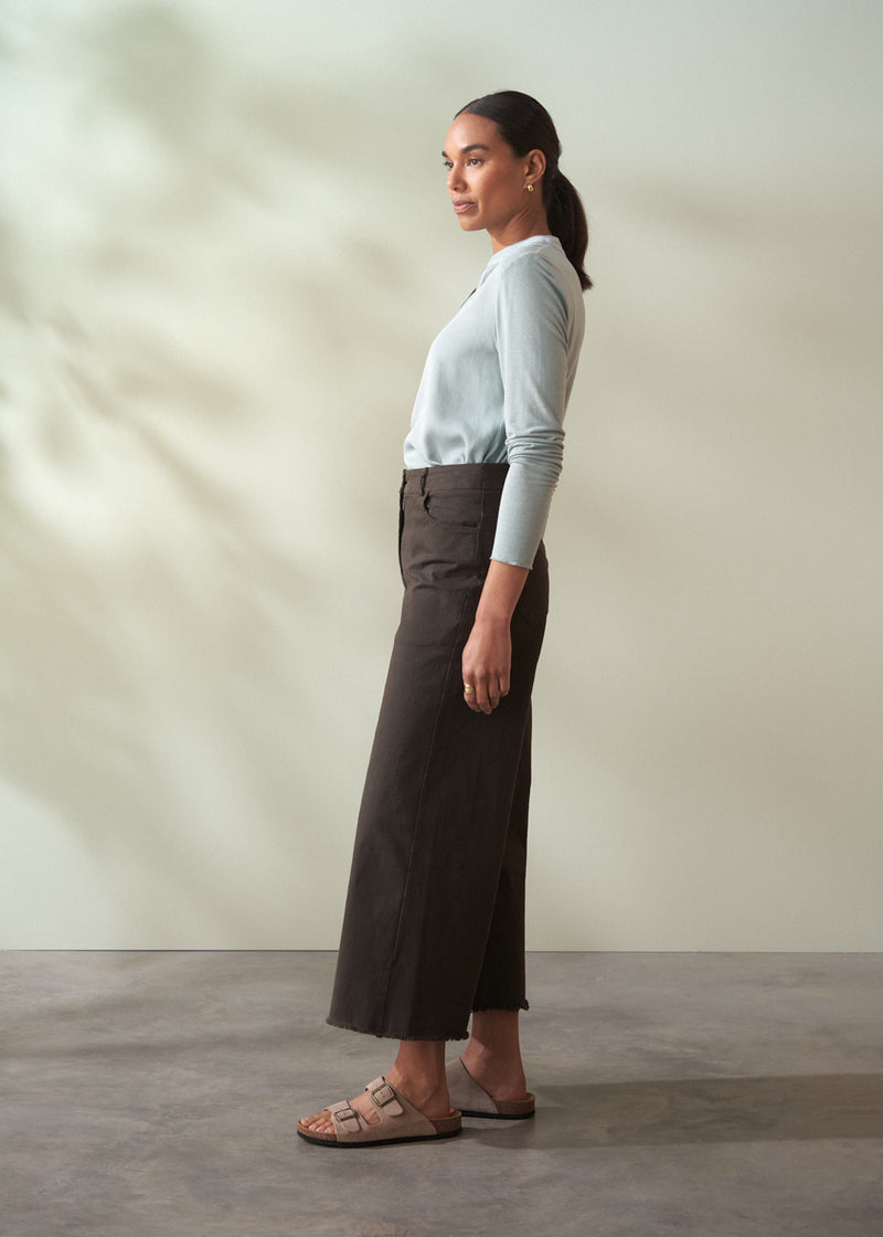 Woman wearing a light gray top and dark brown cropped trousers  standing against a plain background