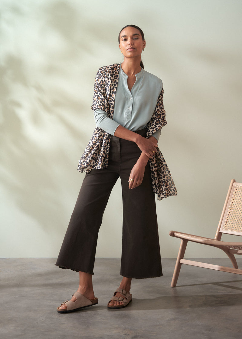 Woman wearing a light blue shirt, dark cropped trousers , and a leopard print scarf in a minimalistic setting.