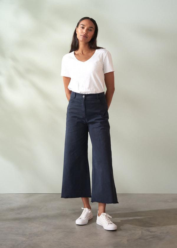 Woman wearing a white t-shirt and dark blue wide leg trousers standing against a plain background