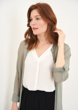 A model wearing a sage green sheer long shirt over a white top, black trousers and black clogs