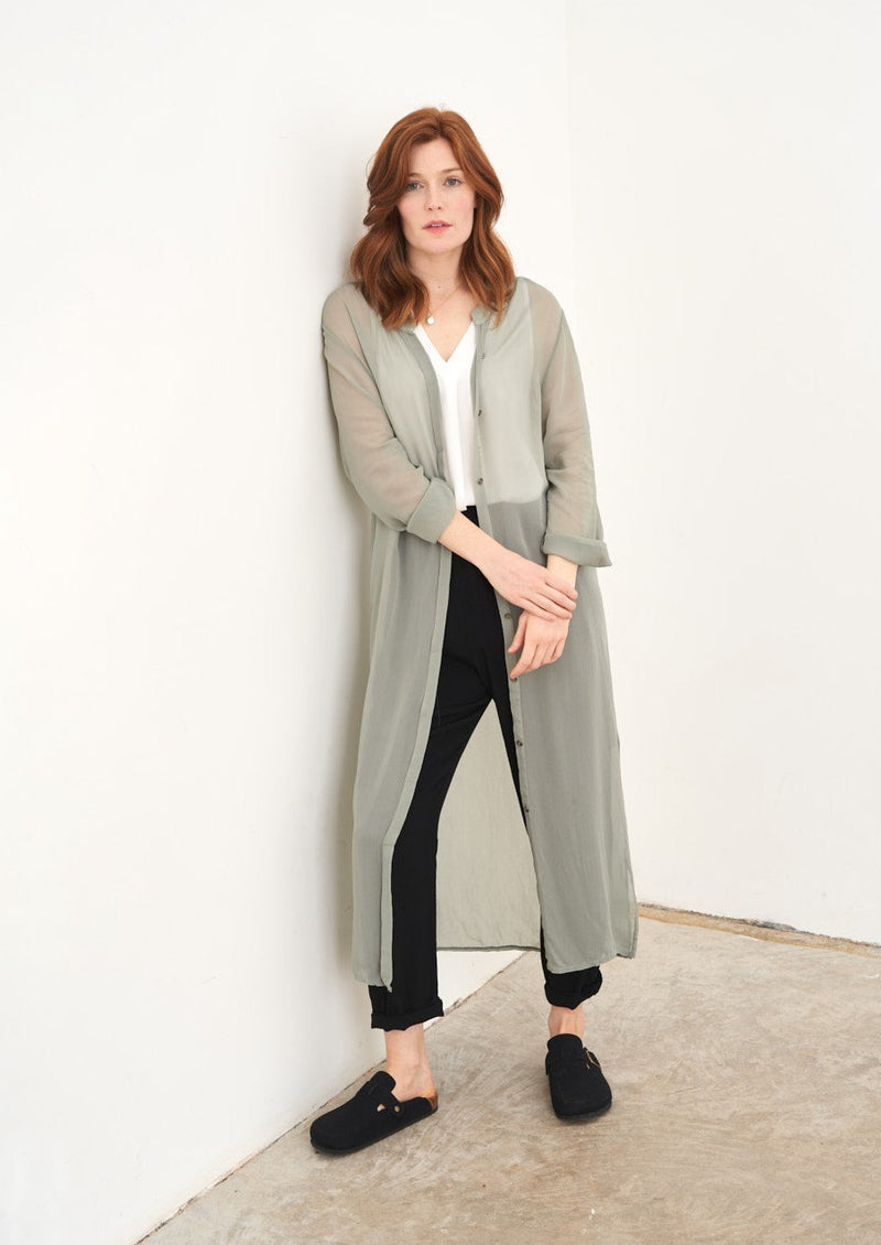 A model wearing a sage green sheer long shirt over a white top, black trousers and black clogs