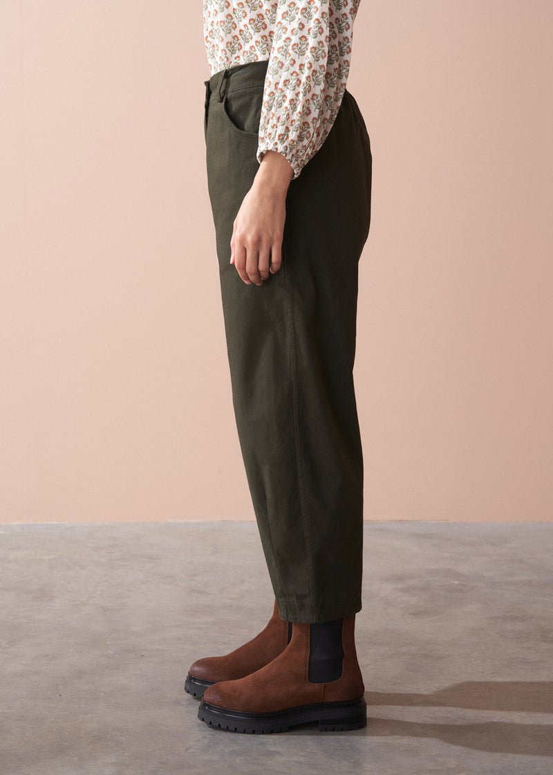 Woman wearing a patterned cotton blouse and green barrel leg trousers against a beige background