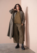 Woman wearing a long coat, sweater, and pants against a beige wall.