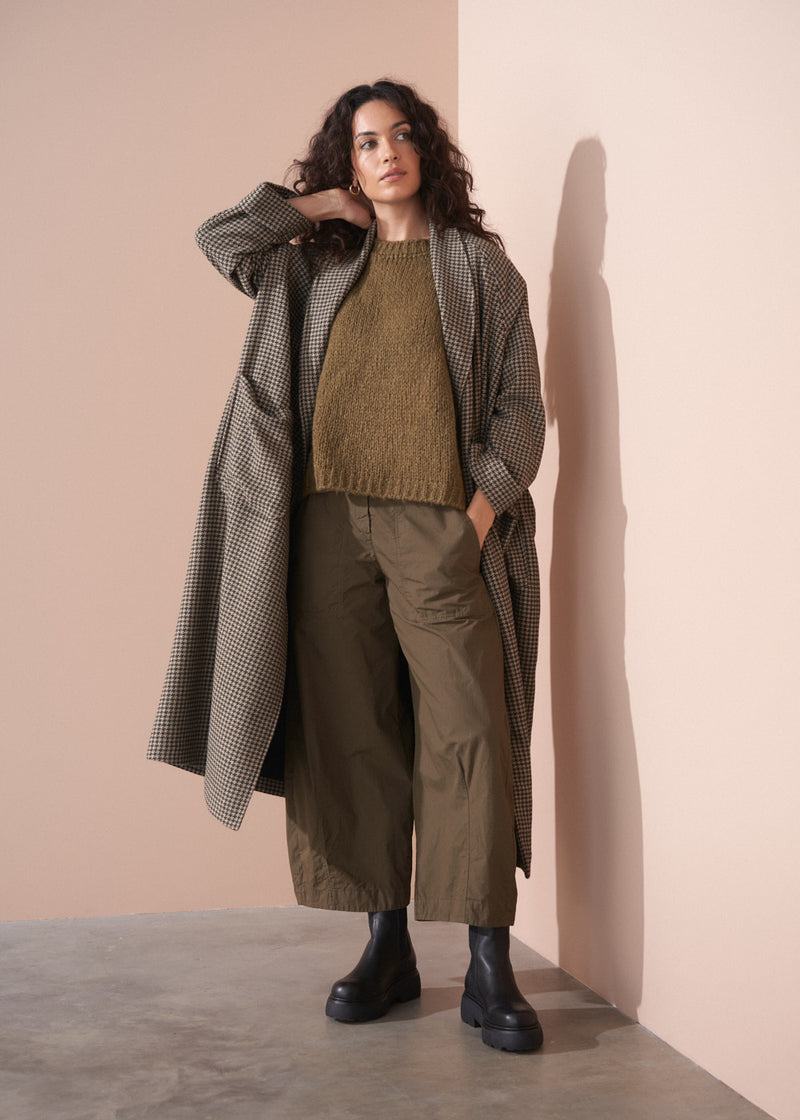 Woman wearing a long coat, sweater, and pants against a beige wall.