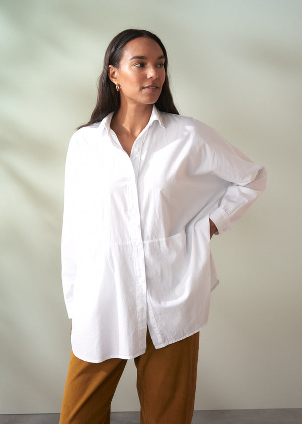 Woman wearing a white oversized smock shirt and brown pants against a plain background