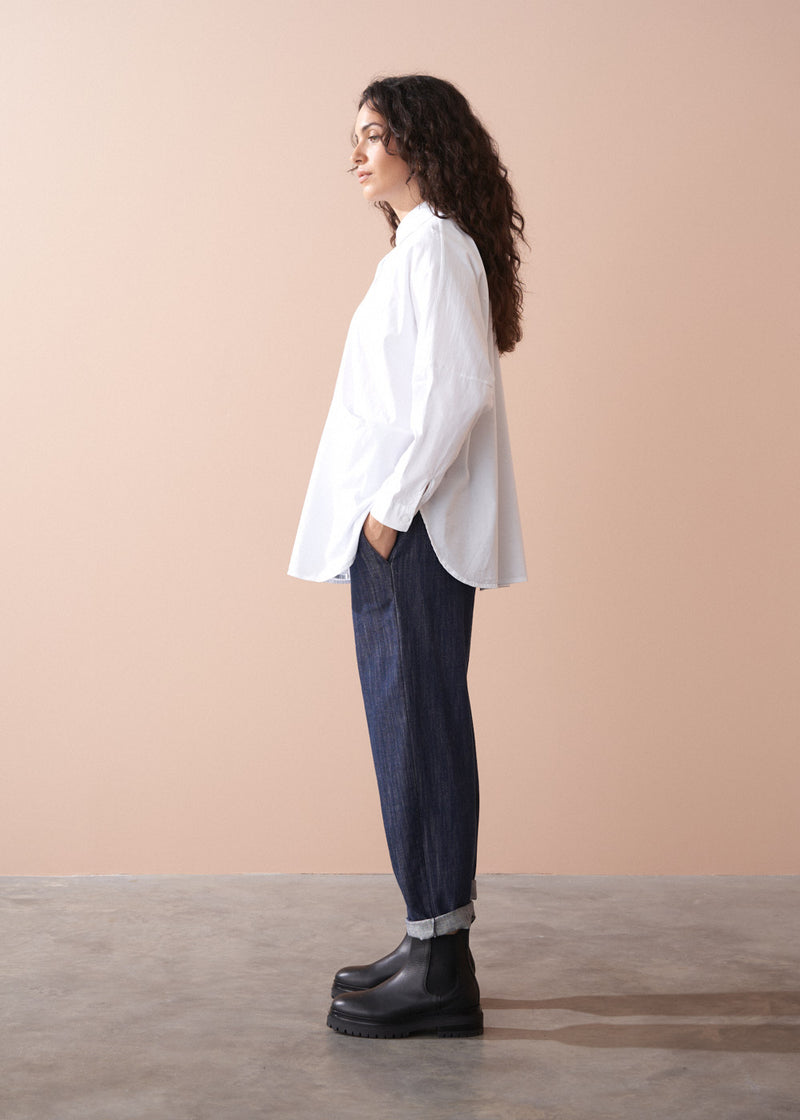 Woman wearing a white shirt, dark jeans, and black boots against a beige background