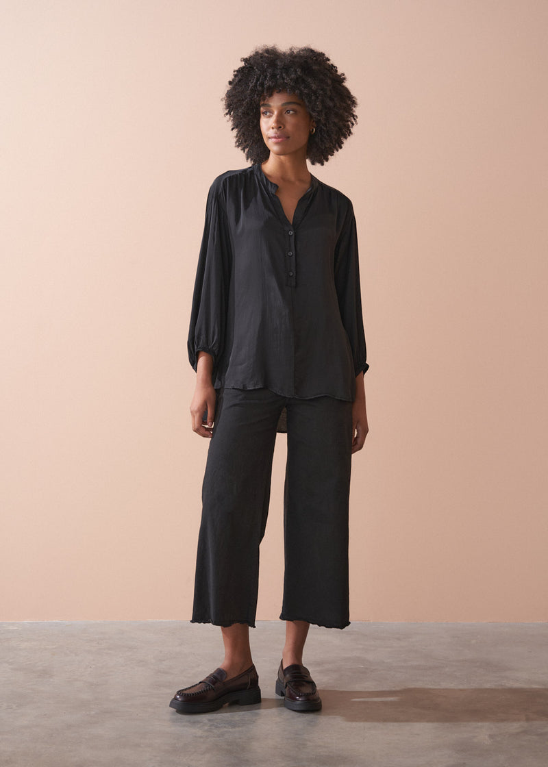 Woman wearing a black satin blouse and black wide leg trousers against a beige background