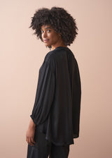 Woman wearing a black satin blouse and black wide leg trousers against a beige background