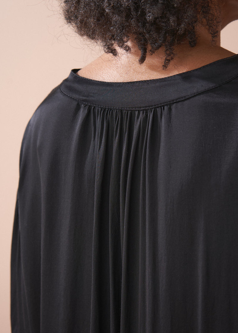 Woman wearing a black satin blouse and black wide leg trousers against a beige background