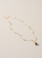 A delicate gold chain necklace with small light blue beads and a teardrop-shaped dark blue pendant, displayed on a textured off-white background.