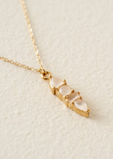 A gold chain necklace with a small vertical pendant featuring three pale pinkish-white teardrop gemstones in a gold setting, photographed on a textured off-white surface.