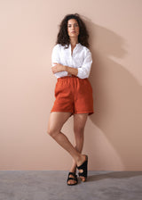 Woman wearing a white shirt and orange shorts against a beige wall.