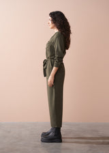 Woman wearing an olive green jumpsuit standing against a beige wall.