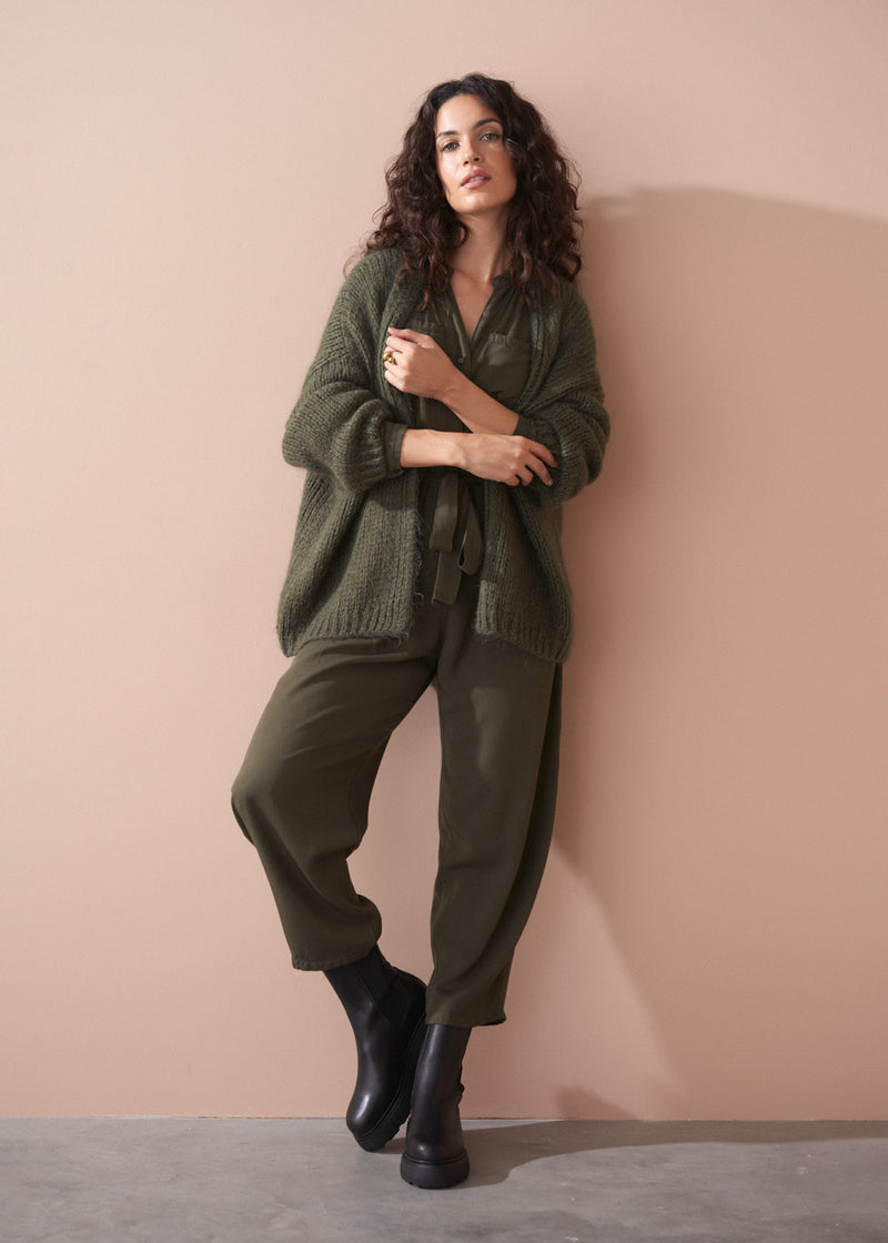Woman wearing a green knitted sweater and jumpsuit against a beige wall.