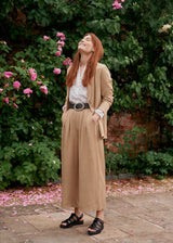 Woman wearing light beige cropped wide leg trousers, matching beige jacket and a white top standing in a garden with flowers.