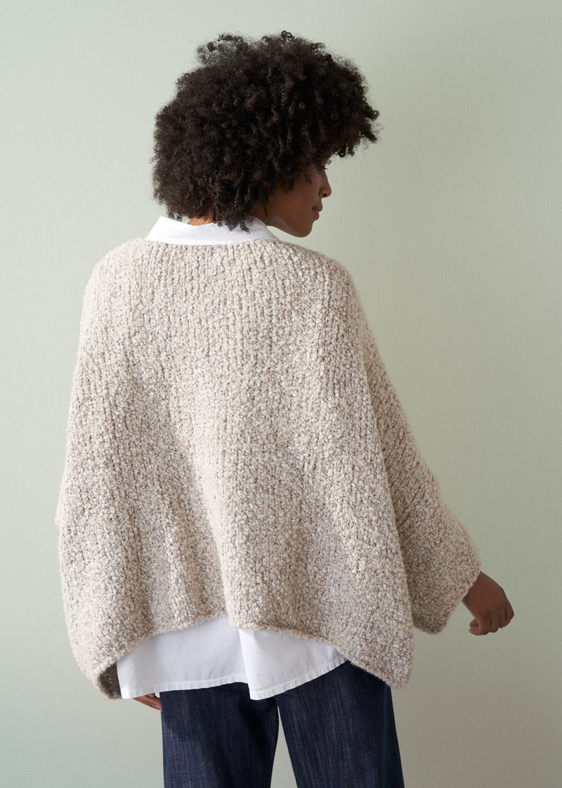 Person wearing an off white boucle sweater and dark blue jeans against a plain background
