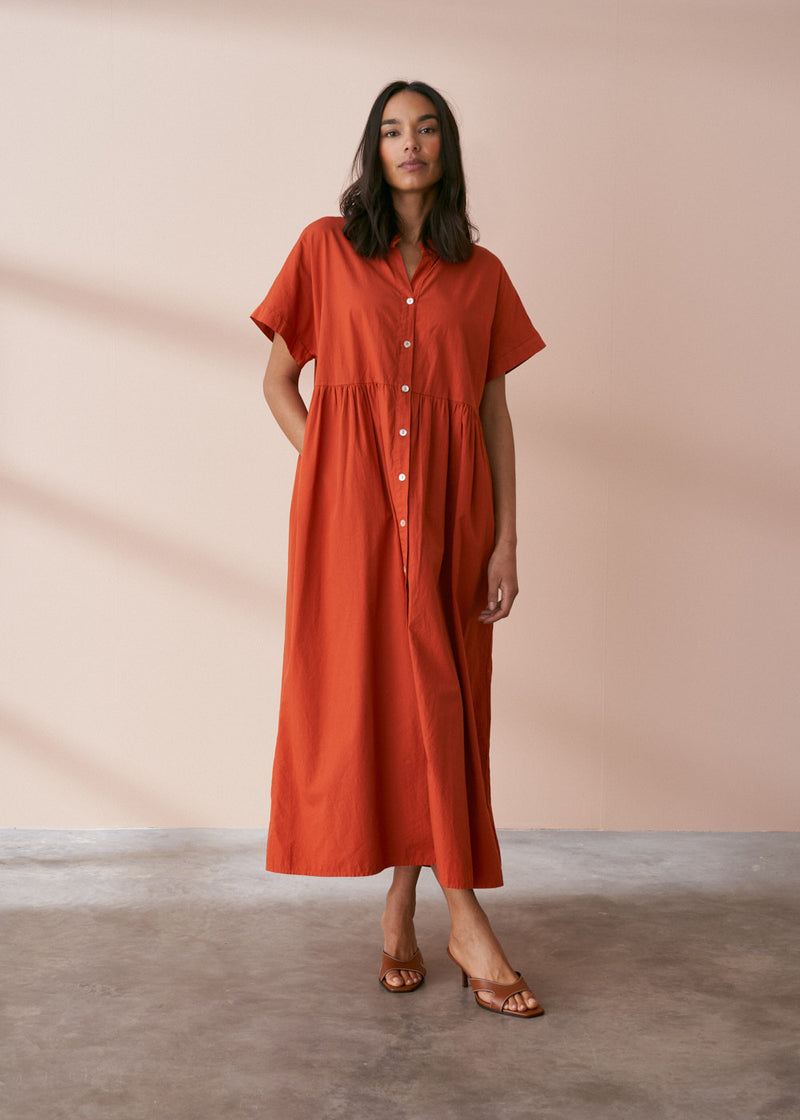 A model wearing a oversized shirt dress in a rusty red tone with mother of pearl buttons