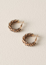 A pair of small gold hoop earrings embellished with rows of round black and clear rhinestones, displayed on a textured off-white background.