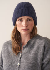 Woman wearing a grey cardigan, black jeans, and a navy wool mix beanie against a plain background