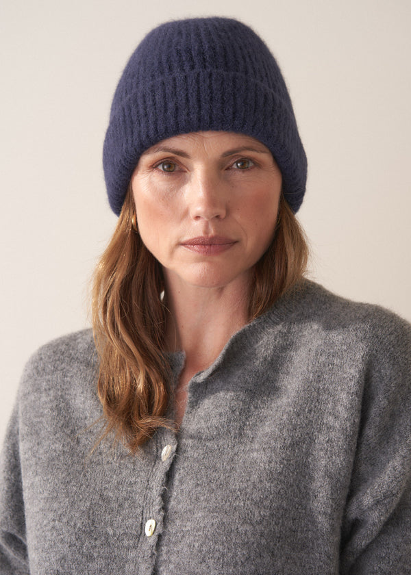 Woman wearing a grey cardigan, black jeans, and a navy wool mix beanie against a plain background