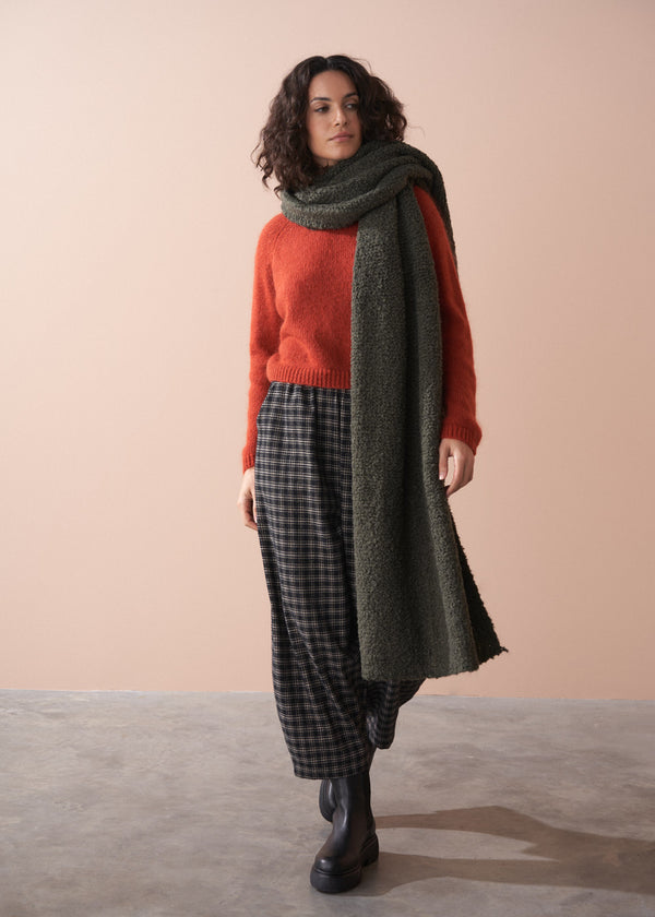 Woman wearing a red sweater, plaid trousers, and green scarf against a beige background