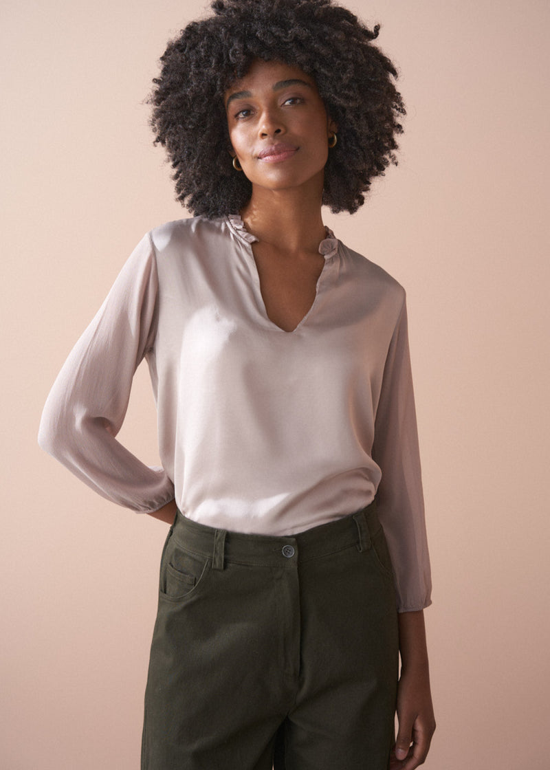 Woman wearing a pale lilac satin  blouse and dark green pants against a beige background