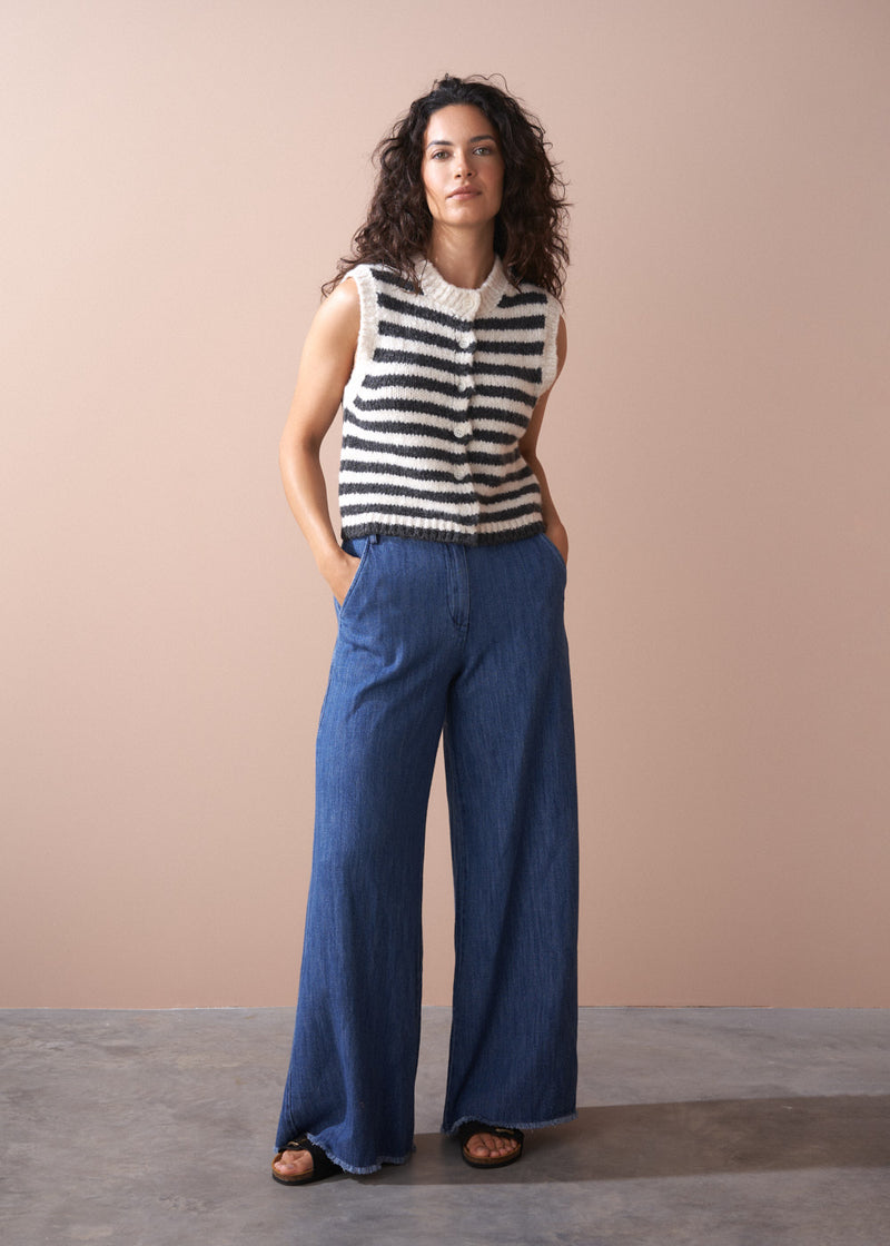 Woman wearing a black and white striped sleeveless sweater and blue wide-leg pants against a beige background