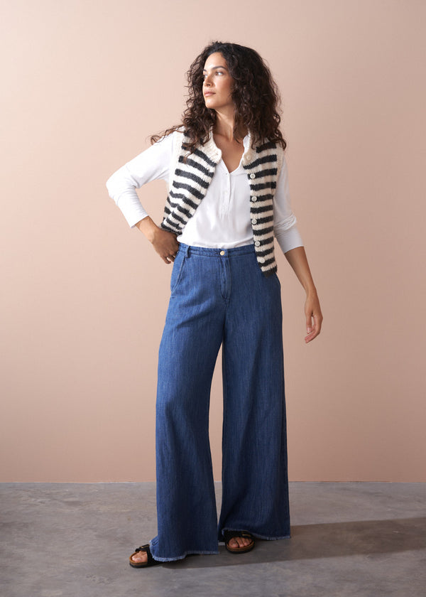 Woman wearing a striped vest, white shirt, and blue jeans against a beige background