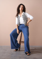 Woman wearing a striped cardigan, white shirt, and blue jeans sitting on a wooden stool against a beige background.