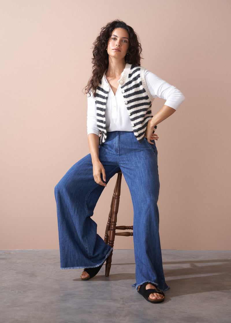 Woman wearing a striped cardigan, white shirt, and blue jeans sitting on a wooden stool against a beige background.